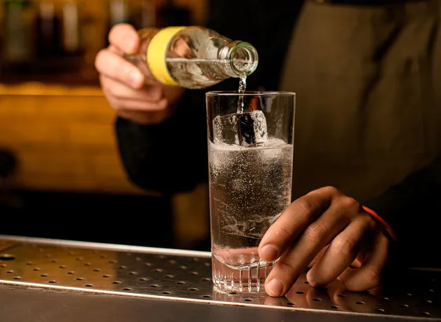 Close-up of glass with cold beverage and ice cubes into which hand of bartender accurate pours fizzy drink from bottle. Cocktail making process.