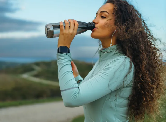 A girl is drinking water from her transparent bottle, a smart watch on her wrist, and headphones in her ears.