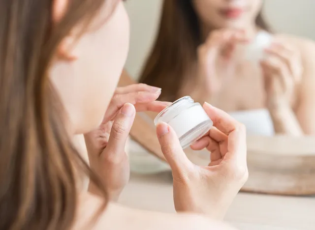 Facial beauty skin care, cute asian young woman, girl in bathrobe reflection into mirror, hand applying moisturizer lotion on her face, holding jar of skin cream before makeup cosmetic routine at home