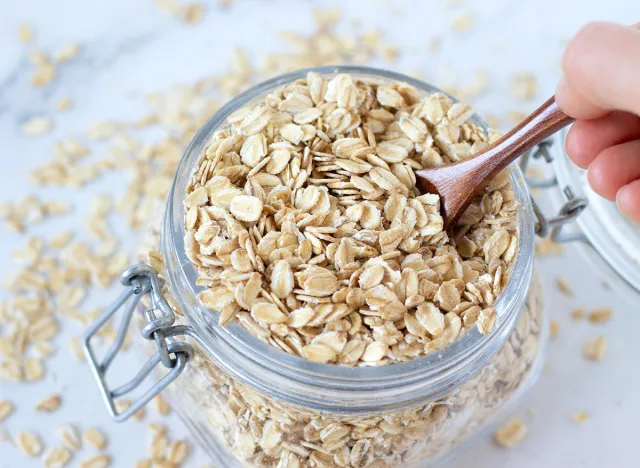 Rolled oats (oatmeal flakes) in a glass jar with hand holding a wooden spoon. Top table view. Organic healthy cereal food for breakfast. Selective focus.