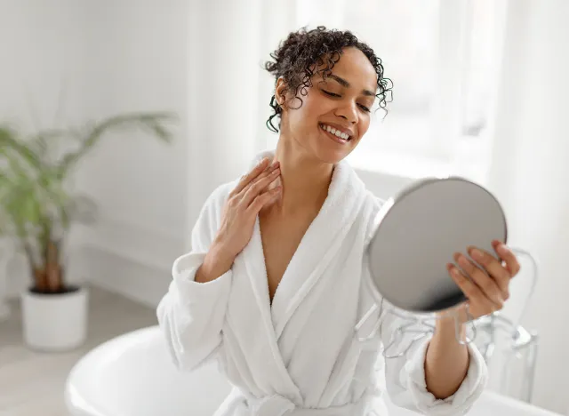 Pretty young black woman in bathrobe looking at mirror and smiling, touching neck and enjoying her moisturised skin, sitting in bathroom, copy space