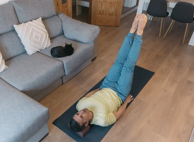 Fit muscular man lies on back on yoga mat in living room, performing abdominal exercises with legs raised. His black cat sleeps on sofa. Home workout.