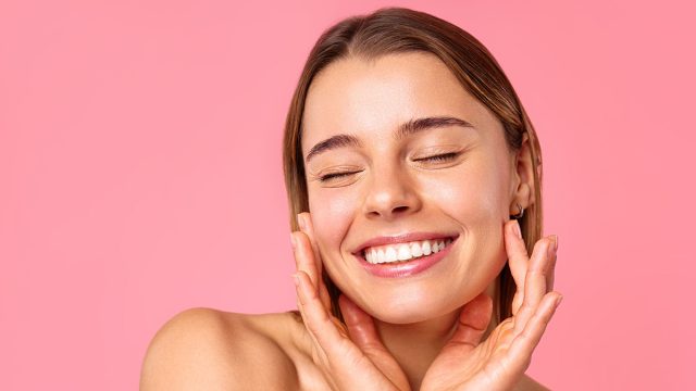 A cheerful young woman flashes a bright smile against a pink backdrop, showcasing her clear, radiant skin and natural beauty. Perfect for wellness, skincare, and positivity concepts.