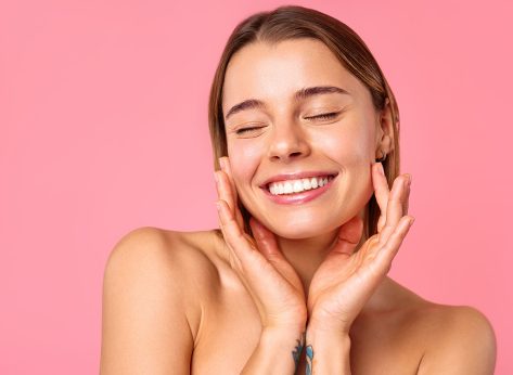 A cheerful young woman flashes a bright smile against a pink backdrop, showcasing her clear, radiant skin and natural beauty. Perfect for wellness, skincare, and positivity concepts.