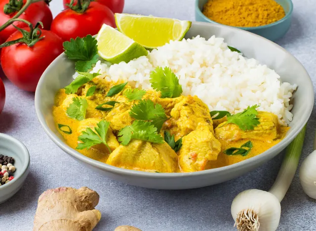 Curry sauce with chicken and rice in a plate, lime, tomatoes, spices and herbs on a light background, horizontal. Traditional Indian dish.