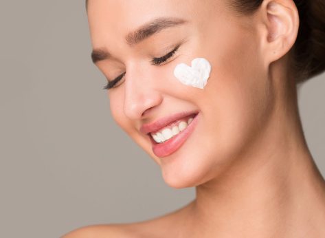 A woman with a radiant smile is shown with a white cream applied on her face. She appears content and relaxed as she enjoys a skincare routine. The cream visibly hydrates and nourishes her skin.