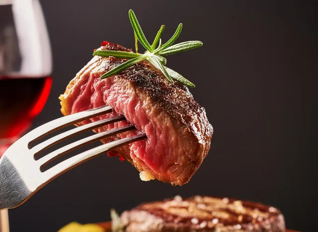 A grilled rib-eye beef steak seasoned with rosemary and accompanied by red wine, all set against a black background. The steak, perched on a fork,is garnished with rosemary and sprinkled with sea Salt