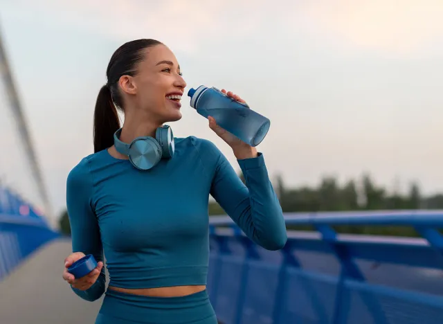 Fit European woman relaxing and drinking water on seaside promenade after running and training outdoors in the evening, panorama, copy space