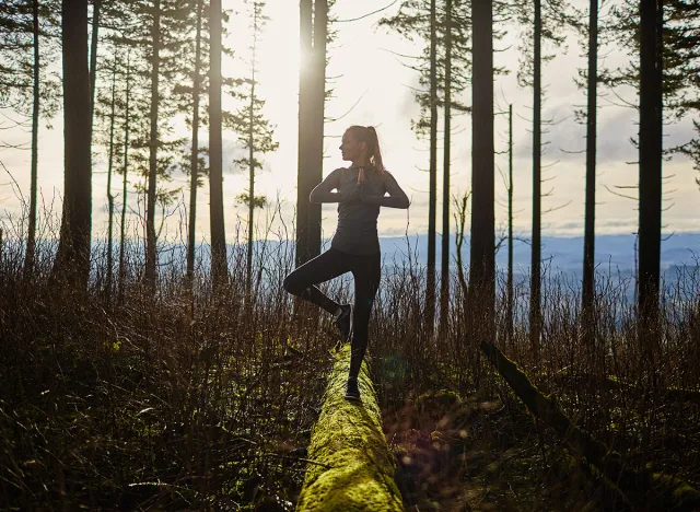 beautiful young girl walking in forest standing on log in yoga tree pose