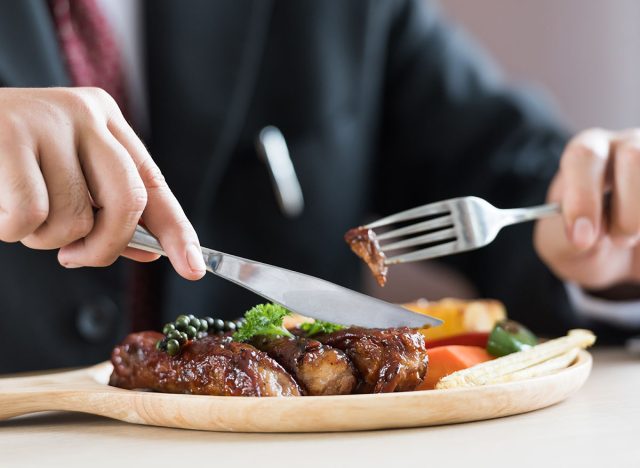 Close up of young businessman eating rib steak on wooden tray at restaurant.