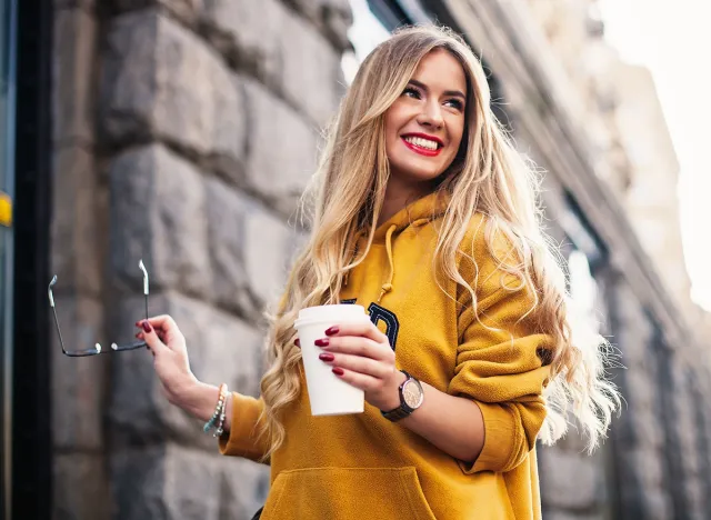 Stylish happy young woman wearing boyfriend jeans, white sneakers bright yellow sweatshirt.She holds coffee to go. portrait of smiling girl in sunglasses Street fashion concept