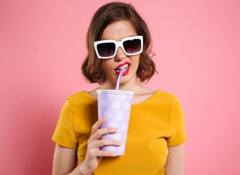 Portrait of a girl in sunglasses holding cup with drink and looking at camera isolated over pink background