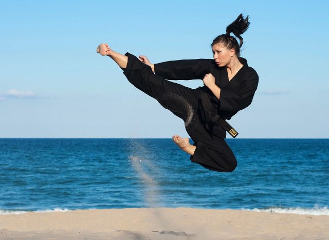 A female, fourth degree, Taekwondo black belt athlete performs a midair jumping kick on the beach.