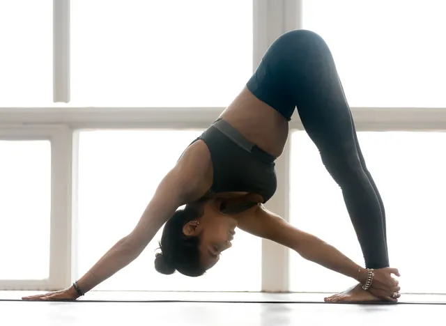 Young sporty woman practicing yoga, doing Revolved Downward Facing Dog exercise, Parivrtta Adho Mukha shvanasana pose, working out wearing sportswear grey pants, top, indoor full length at yoga studio