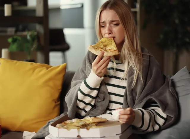 Portrait of sad young blond woman dealing with stress by eating food sitting on sofa at home Upset young woman wrapped in blanket crying and eating pizza indoors. Emotional, Eating