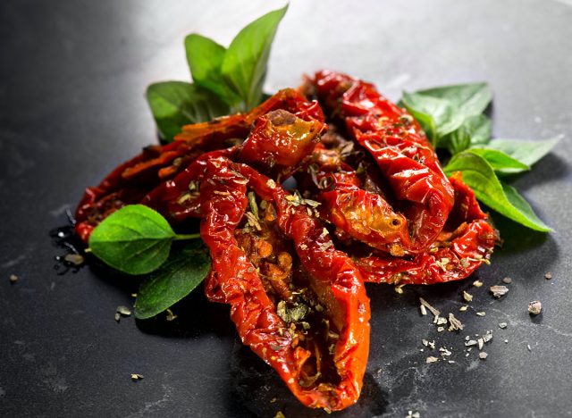 Sun dried tomatoes with aromatic herbs, oregano, marjoram leaf. Over black stone background, close up. Italian cuisine, healthy vegan food, Mediterranean diet. Cured, Dry tomato