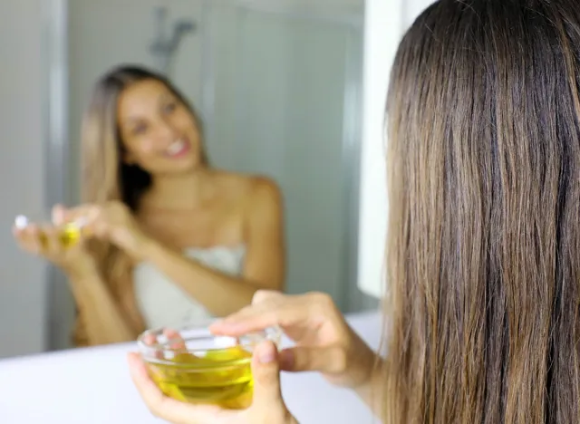 Smiling girl applying oil mask on hair in front of a mirror. Hair care concept. Focus on hair.