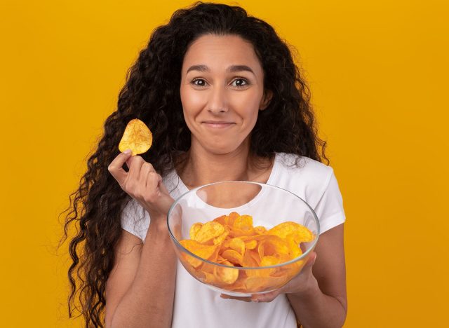 Tasty Fast Food Concept. Portrait Of Smiling Cute Young Lady Enjoying Delicious Potato Crisps Holding Glass Bowl, Posing With Chips In Hand Standing Isolated On Yellow Orange Studio Background Wall