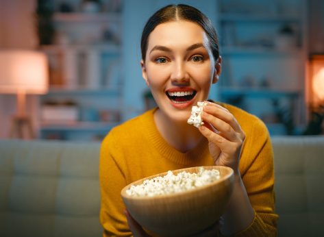 Young woman watching projector, TV, movies with popcorn in the evening. Girl spending time at home.