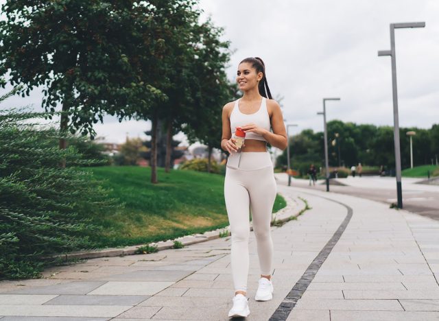 Full length of happy female athlete in sportswear smiling and looking away while walking on paved sidewalk with bottle in hands on street