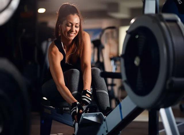 Happy female athlete adjusting rowing machine and listening music on earphones during sports training in a gym.