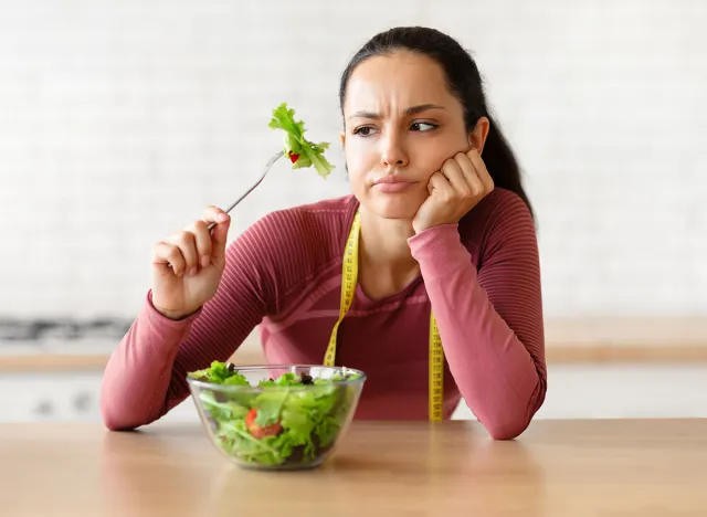 Unhappy young fitness lady in fitwear eating vegetable salad posing with fork at kitchen table indoor, struggling from lack of appetite, tired of diet menu and low carbs dishes. Dieting issues