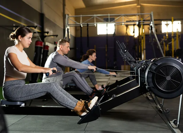 Girl performs exercises with rowing machine. Female gym visitor performs series of repetitions movements on simulator. Shot of sexy muscular woman in sportswear and good physique on grey background