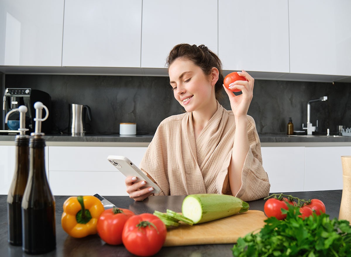 Portrait of young woman ordering groceries on smartphone app, holding tomato, sitting near chopping board with vegetables. Girl looking for recipe online, using mobile phone.