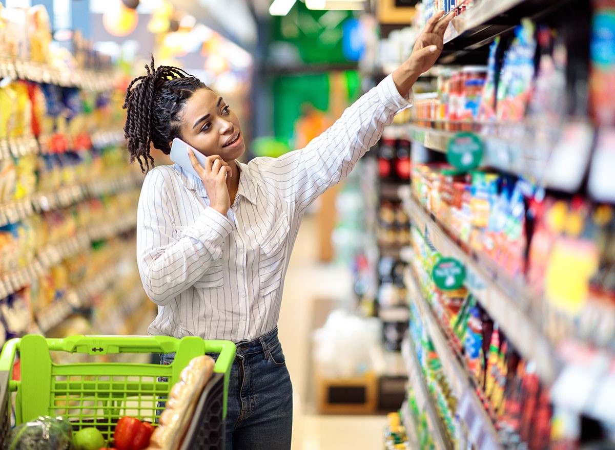 African American Woman Talking On Mobile Phone Buying Food During Grocery Shopping In Supermarket Store. Black Female Customer Choosing Groceries Chatting On Cellphone In Shop.