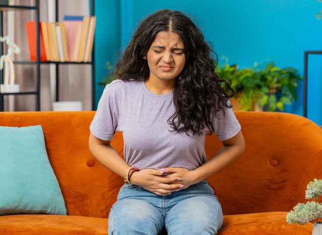 Sick ill Indian woman suffering from period cramps, painful stomach ache on sofa at home room. Girl holding belly, feeling abdominal menstrual pain. Abdominal pain, gastritis, diarrhea, indigestion