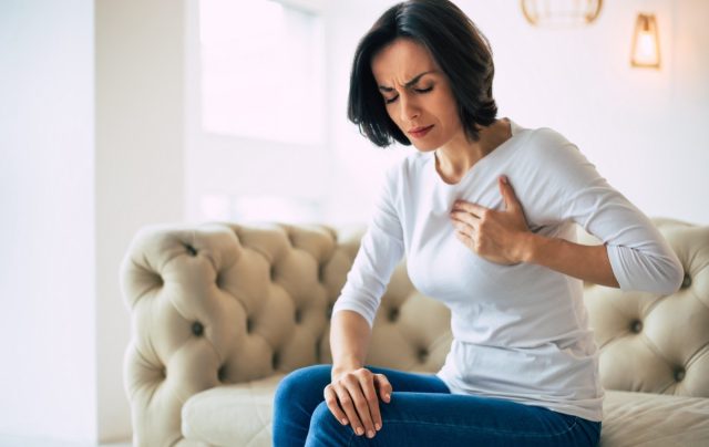 Close-up photo of a brunette woman who is sitting on a couch with her eyes closed and her left hand touching her heart area.