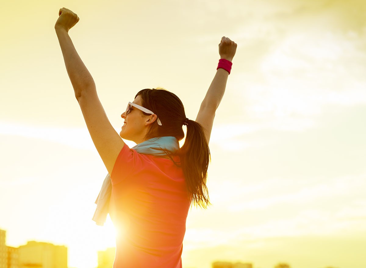 Happy successful sportswoman raising arms to the sky on golden back lighting sunset summer. Fitness athlete with arms up celebrating goals after sport exercising and working out outdoors. Copy space. sunrise, sunset