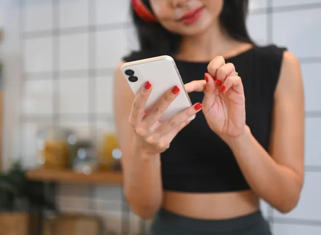 Happy young fitness woman listening music through headphones and using mobile phone in kitchen. iPhone, mobile, kitchen