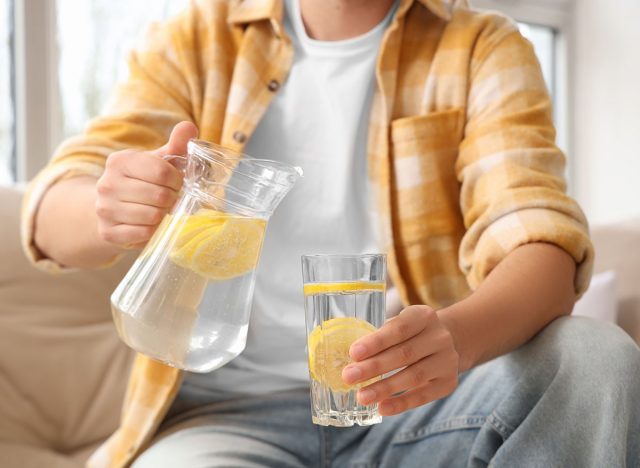 Young man with jug and glass of lemon infused water sitting on sofa at home, closeup