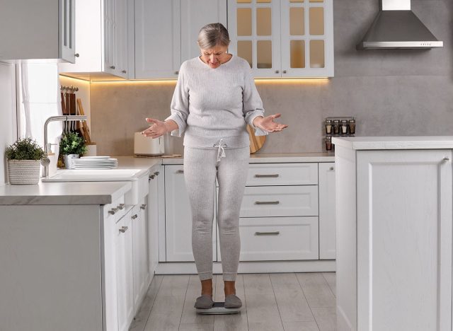 Menopause, weight gain. Concerned woman standing on floor scales in kitchen