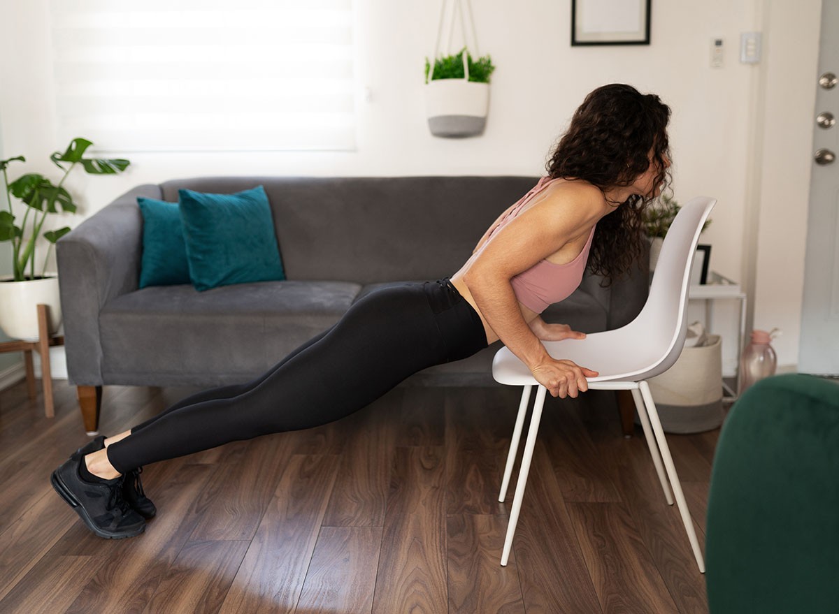 Side view of an active young woman wearing sportswear doing push ups with the help of a chair at home