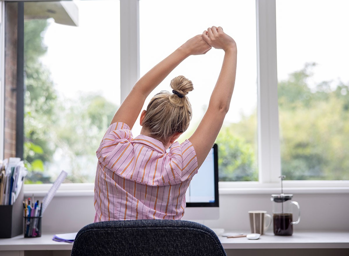 Rear View Of Woman Working From Home On Computer In Home Office Stretching At Desk