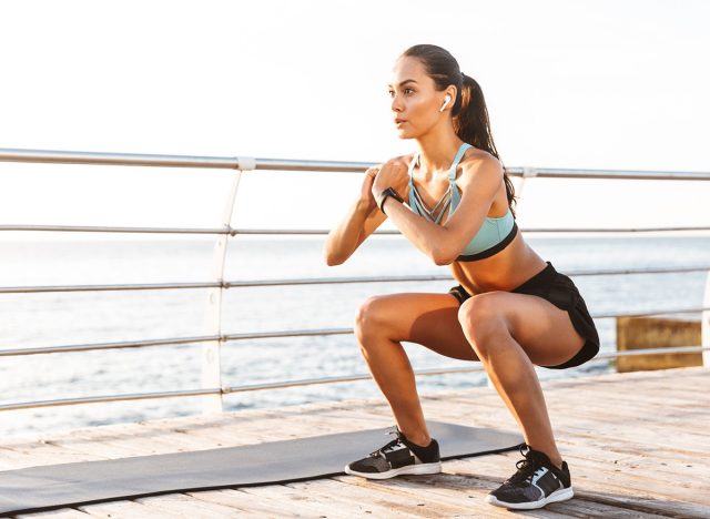Photo of attractive concentrated woman 20s in tracksuit squatting near sport mat during workout on boardwalk at seaside