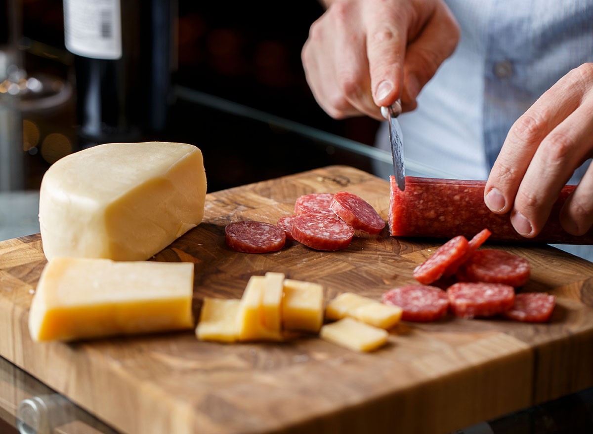 hands slicing salami and cheese on a table with wine