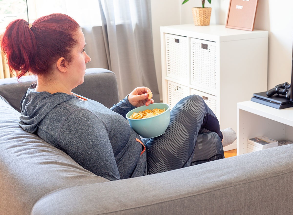 Middle age woman relaxing on the sofa, watching TV and eating chips