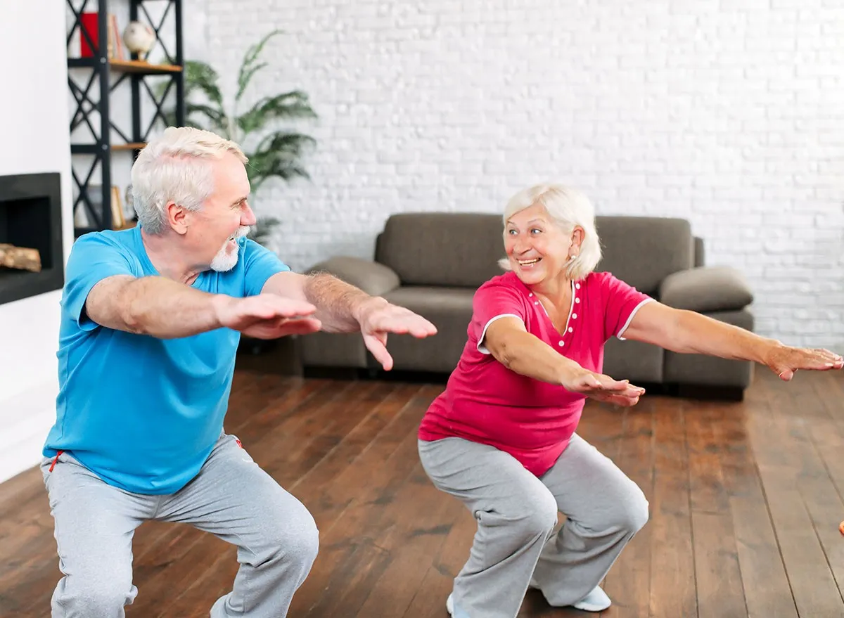 Elderly couple doing squats together at home. Cheerful grey-haired spouses doing exercises, look to each other and smiles, mature
