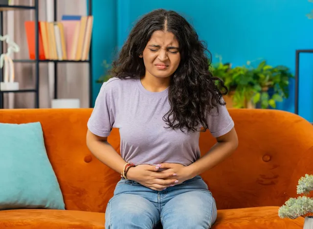 Sick ill Indian woman suffering from period cramps, painful stomach ache on sofa at home room. Girl holding belly, feeling abdominal menstrual pain. Abdominal pain, gastritis, diarrhea, indigestion