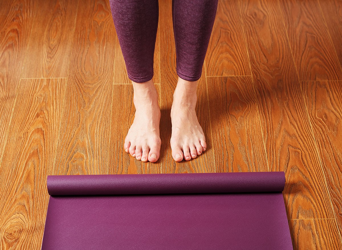The girl's feet stand in front of an unfolded Yoga mat on the wooden floor. Yoga practice classes, asana exercises