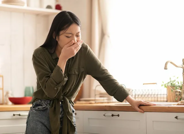 Young asian woman in casual feeling nausea, cover mouth, standing next to table in kitchen, copy space. Toxicosis during pregnancy, food poisoning concept