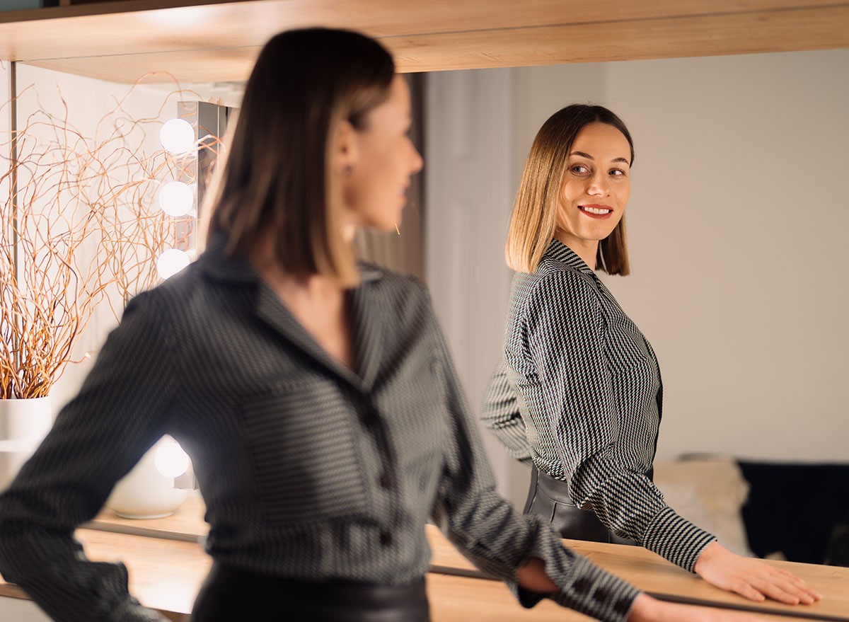 Self-confident Woman looking at her reflection into the mirror indoors. Beautiful interior design