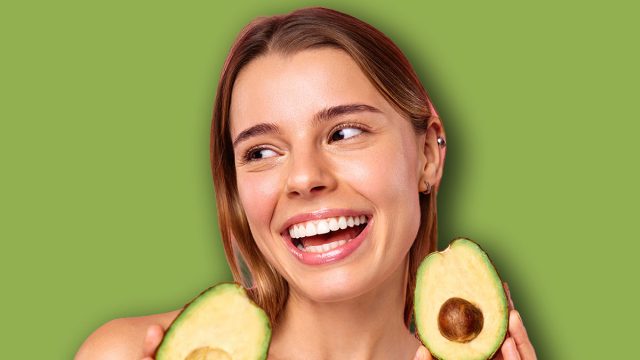 A young woman with a tattoo poses happily with an avocado on a pink background, symbolizing wellness, beauty, skincare, nutrition, and a positive lifestyle. Her smile and appearance exude happiness