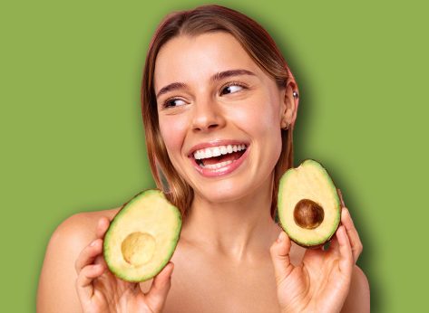 A young woman with a tattoo poses happily with an avocado on a pink background, symbolizing wellness, beauty, skincare, nutrition, and a positive lifestyle. Her smile and appearance exude happiness