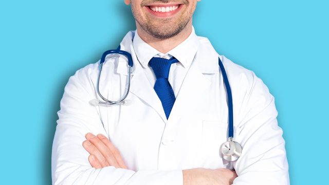 Young male doctor standing confidently with crossed arms wearing a white coat and stethoscope on a bright blue background