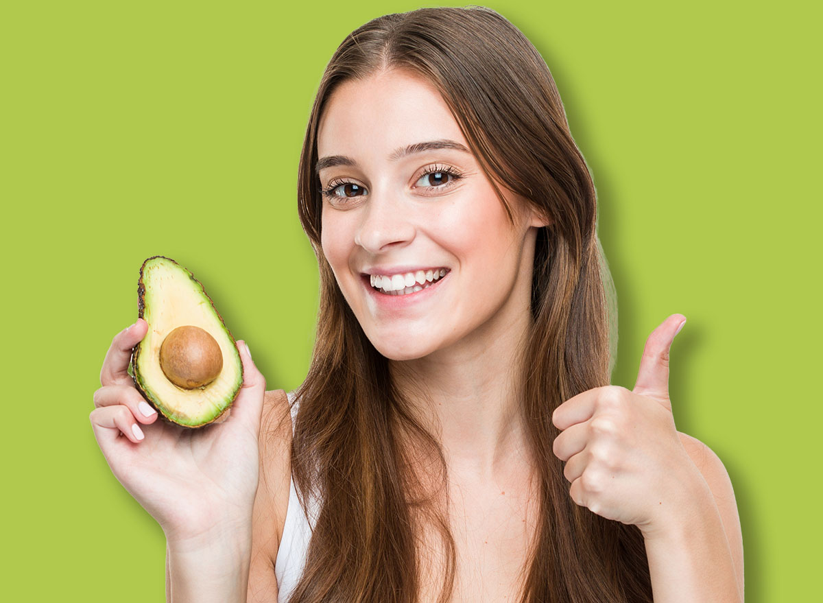 Young caucasian woman holding an avocado smiling and raising thumb up