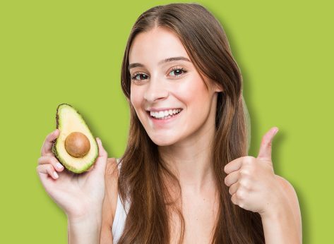 Young caucasian woman holding an avocado smiling and raising thumb up